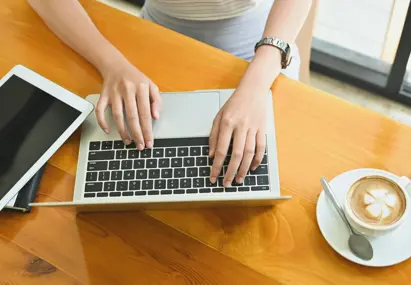 Handen typen op laptop aan houten tafel, met tablet, notitieboek en cappuccino ernaast in een lichte werkruimte.