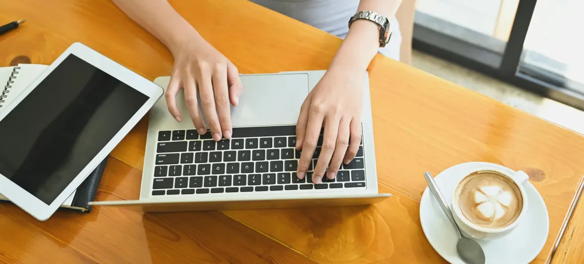 Handen typen op laptop aan houten tafel, met tablet, notitieboek en cappuccino ernaast in een lichte werkruimte.