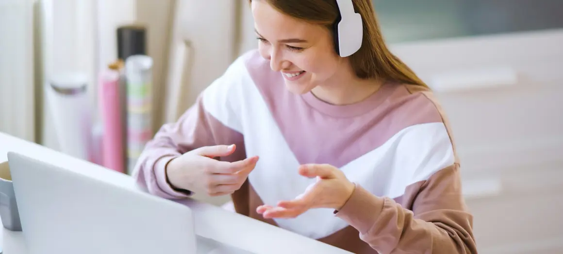 Vrouw met koptelefoon voert online gesprek via laptop aan bureau, glimlachend en gebarend tijdens videogesprek.
