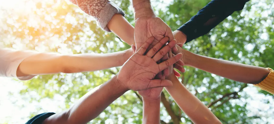 Meerdere handen van diverse mensen op elkaar gelegd in de buitenlucht, symbool voor samenwerking en verbinding.