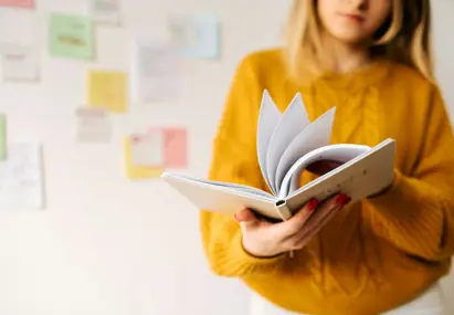 Vrouw in gele trui bladert door notitieboek, met kleurrijke post-its op de muur op de achtergrond