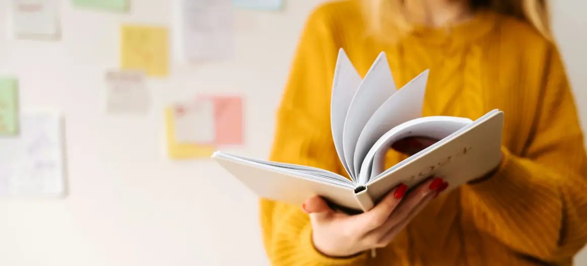 Vrouw in gele trui bladert door notitieboek, met kleurrijke post-its op de muur op de achtergrond