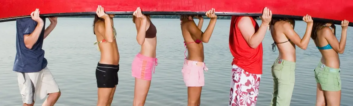 Groep mannen en vrouwen in badkleding houden een omgekeerde kano vast die hun hoofden verbergt