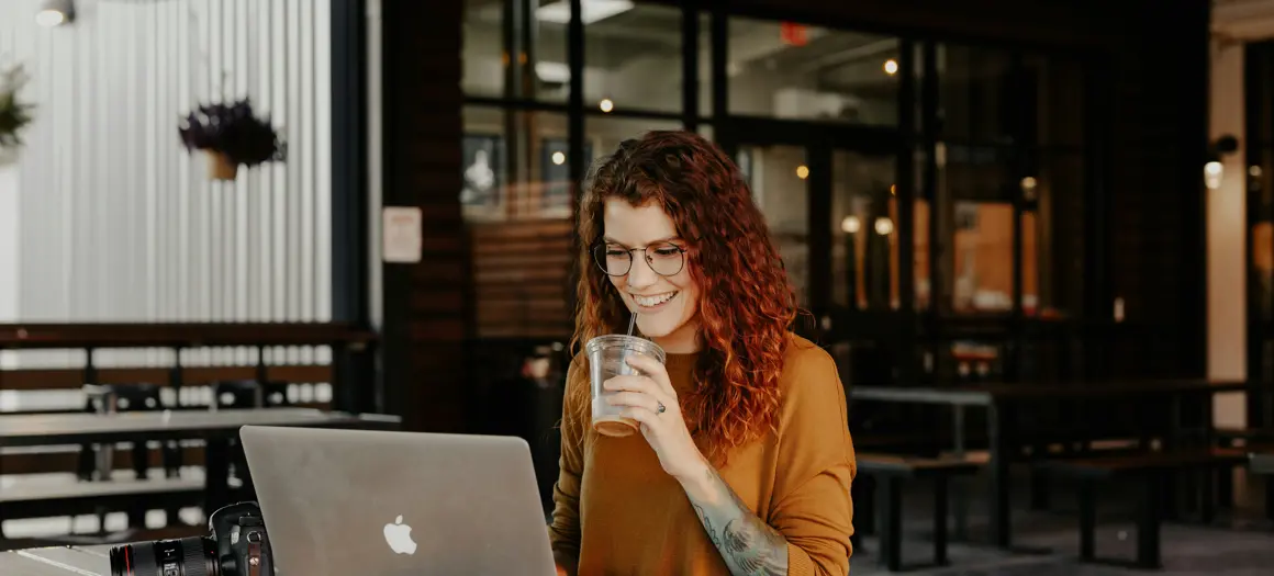 Vrouw aan tafel achter laptop met drankje in haar hand